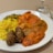 A plate of Indian cuisine featuring vibrant yellow rice with peas, assorted curries with rich red and brown gravies, and two brown kebabs. The dish is served on a white plate alongside cutlery on a neutral tablecloth background.